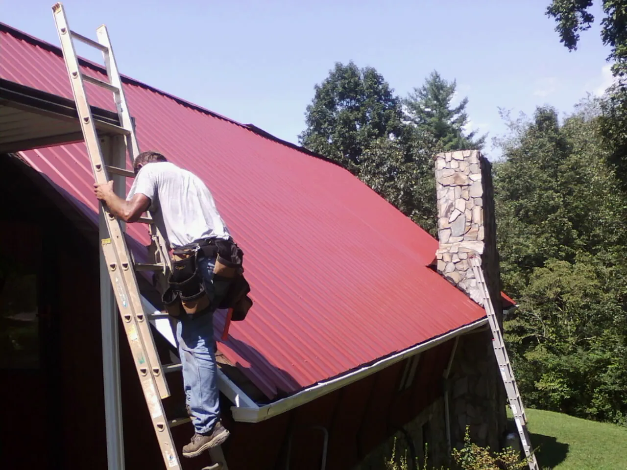 Roofer on a ladder beside a red metal roof and stone chimney — Soffit & Fascia Repair by Roofing Brothers in Sperry